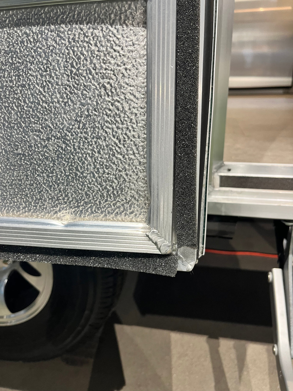 Close-up of the corner of a metal panel with a textured surface, featuring Airstream Foam Tape for Screen Door (Part #365013-01) and an aluminum frame—likely part of an Airstream screen door—positioned next to a vehicle tire.