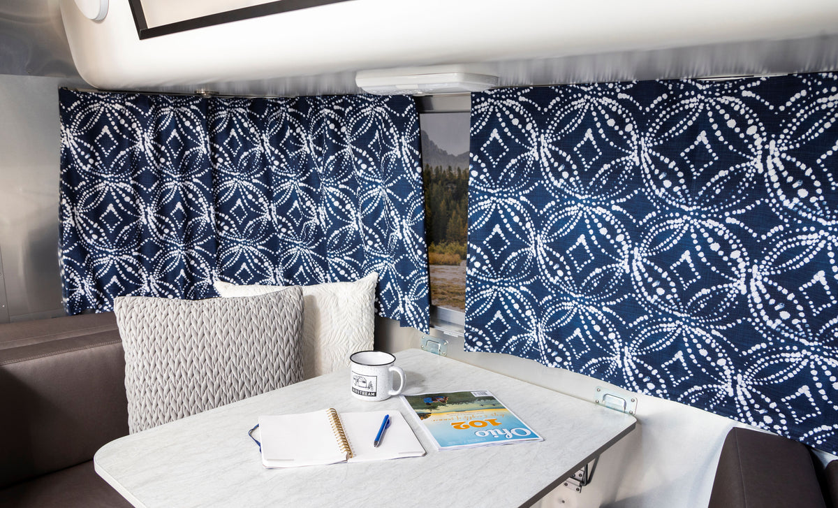 A cozy RV dinette featuring Airstream Printed Custom Curtains for Pottery Barn Travel Trailers, with a table set with an open notebook, pen, coffee mug, and an Ohio travel book. Cushioned benches and pillows surround the table.