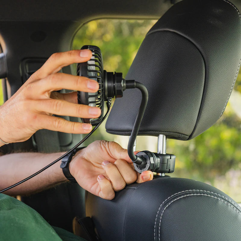 A person attaches the USB Powered Camping Fan by Luno to the metal rods of a car’s headrest using a clamp, positioning the fan towards the backseat—an ideal setup for car camping or van life enthusiasts who want extra comfort while traveling.