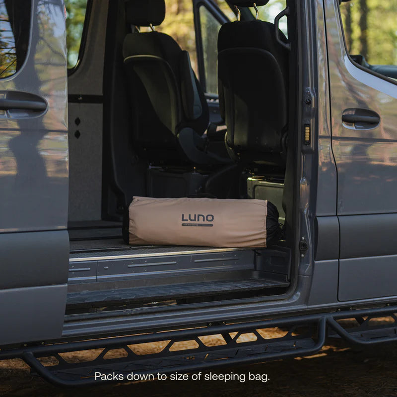 A Tan Luno bag is packed down and placed on the floor inside a gray van with the sliding door open; caption reads, Packs down to the size of a sleeping bag—perfect for your Touring Coach Front Cab Air Mattress by Luno or other van sleeping solutions.