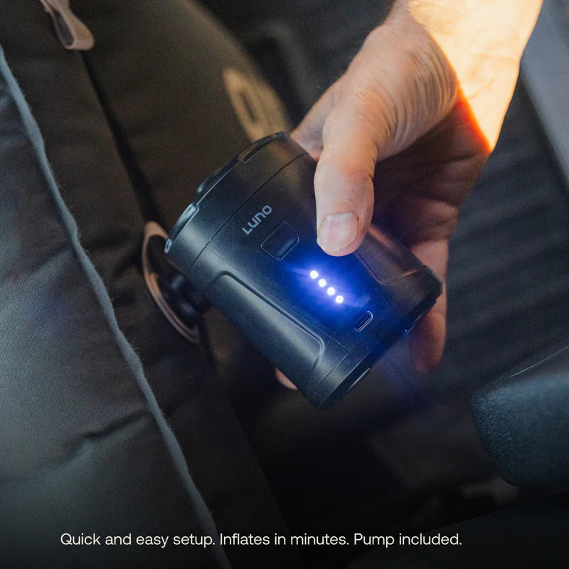 A hand holds a compact black air pump with blue LED lights, next to a car seat—perfect for inflating your Touring Coach Front Cab Air Mattress by Luno. Text at the bottom reads: Quick and easy setup. Inflates in minutes. Pump included.