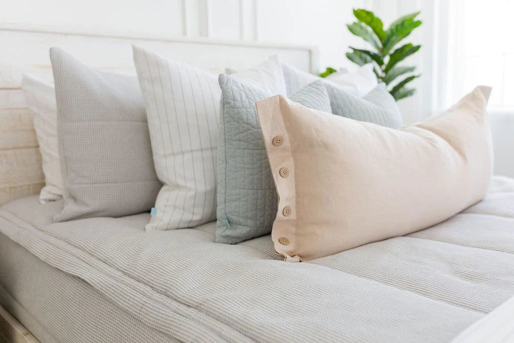A neatly made bed features a row of decorative pillows in neutral tones, including the Harper Medium Pillow Cover by Beddy’s—a long beige accent pillow cover with buttons—set against a white background with a green potted plant in the corner.