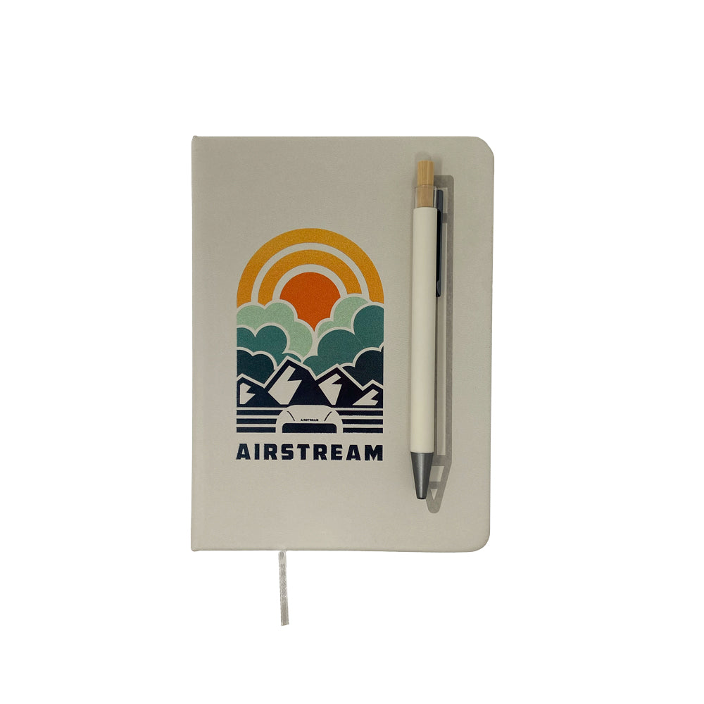 The Airstream Horizons Journal and Pen Set showcases a white journal with a vibrant geometric mountain and sun design labeled “AIRSTREAM,” accompanied by a matching white pen clipped to the cover—an ideal travel companion, presented on a plain white background.