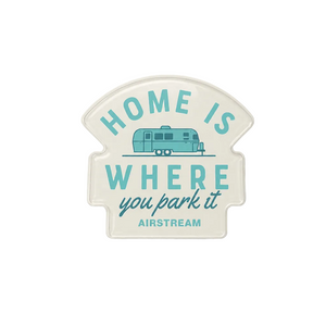 A die-cut vinyl sign with a vintage-style cream background and turquoise lettering that reads 'HOME IS WHERE you park it' with 'AIRSTREAM' at the bottom, featuring a small turquoise retro camper illustration. The sign is photographed against a neutral light gray background.