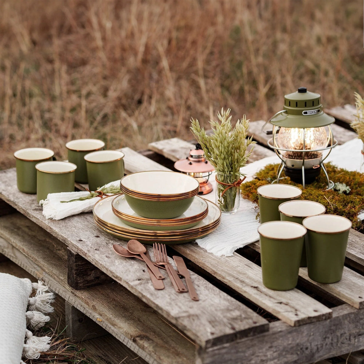 Create a rustic outdoor table setting on wooden pallets with Barebones Enamel Dining Sets—featuring green enamelware cups, plates, and bowls—paired with bronze cutlery, a lantern, small plants, and a white cloth. This scene in a grassy field is perfect for capturing the charm of an Airstream galley under the open sky.