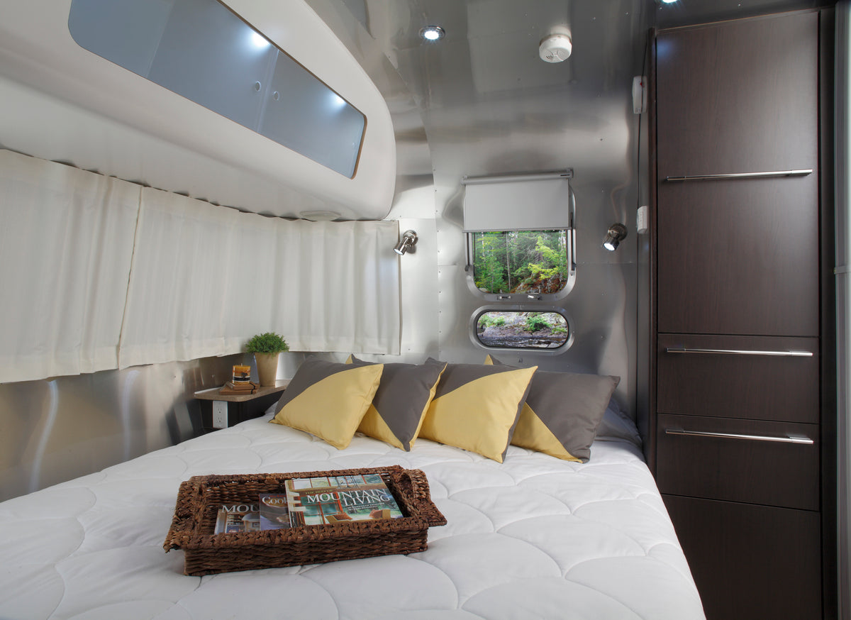 Interior of a modern RV featuring a neatly made bed with four yellow and gray pillows, a wicker tray with magazines, small window, potted plant, dark wood cabinets, and Airstream Solid Custom Curtains for International Signature Travel Trailers.