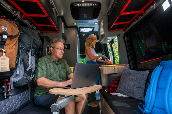 A man sits inside a camper van using a laptop on a fold-out table, with a woman working in the kitchen area behind him. The interior features dark gray seating, gray pillows, a blue backpack on the right, and red trim on the ceiling.