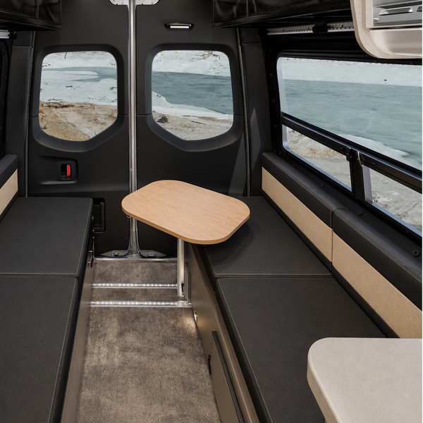 The interior of a camper van or RV showing a spacious seating area with two long dark gray bench seats on either side of a central aisle, a small fold-out wooden table, and windows looking out onto a beach and ocean view.