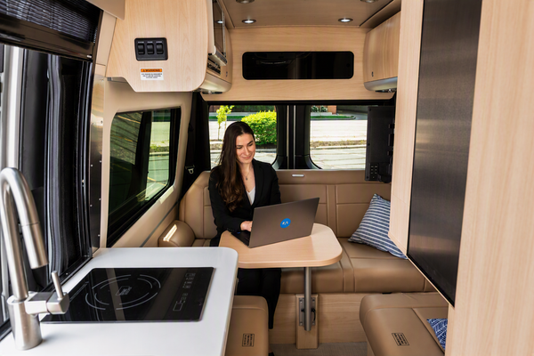 A woman sits inside a recreational vehicle or camper, working on a laptop at a small table. The interior features light wood paneling, beige leather seating, a built-in kitchenette with a sink and cooktop, and large windows.