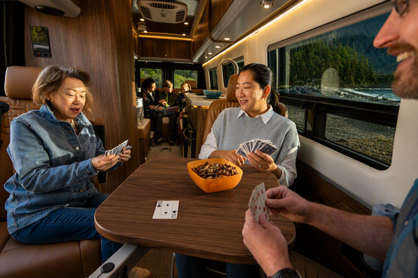 Three people sitting inside a camper trailer or RV, playing cards at a small wooden table with an orange bowl of snacks in the center. The interior features wood paneling and a window showing a mountain landscape outside.