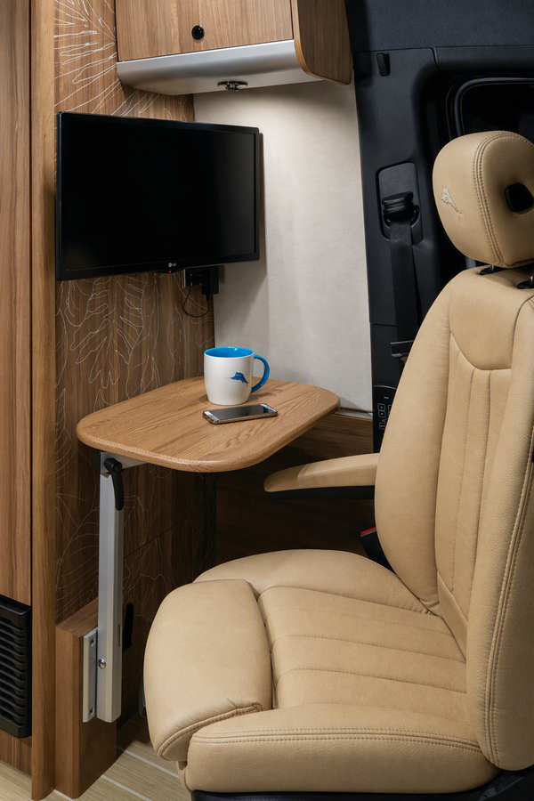 Beige leather recreational vehicle cab seat with a fold-out wooden side table, wall-mounted TV, and a blue and white mug on a phone inside the cab.