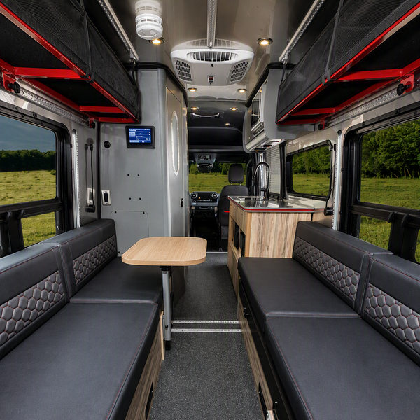 The interior of a camper van or RV kitchen shown from the back, featuring two long black padded bench seats on both sides, a small wooden table in the center, and a wood-and-black kitchenette unit at the rear.