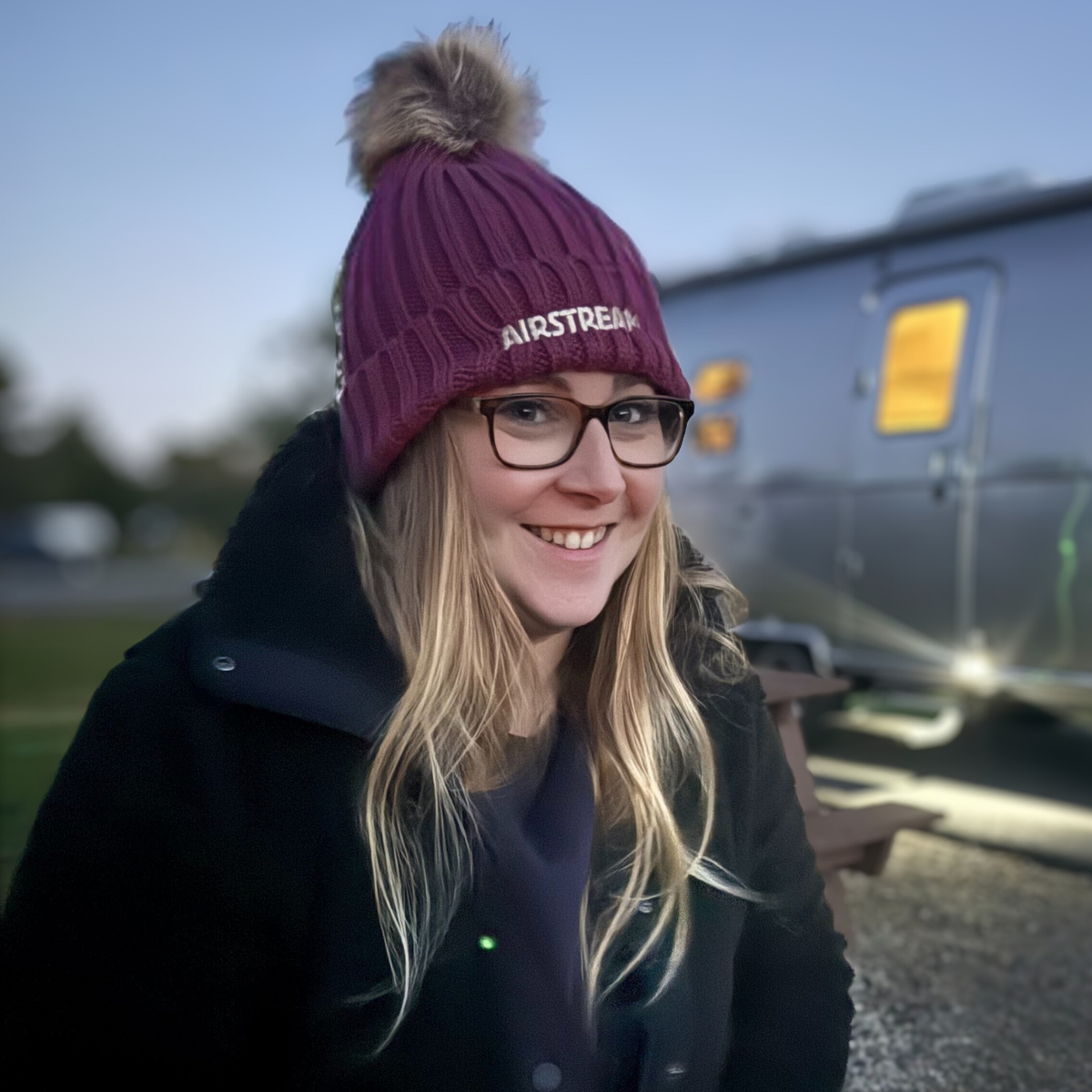 A smiling woman with long blonde hair and glasses wears the Airstream Ribbed Pom Beanie in maroon, standing outdoors beside a silver trailer at dusk.