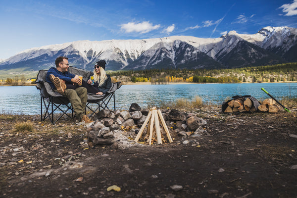 A couple relaxes in black camping chairs beside a campfire ring at a lakeside, with snow-capped mountains and a blue sky in the background.