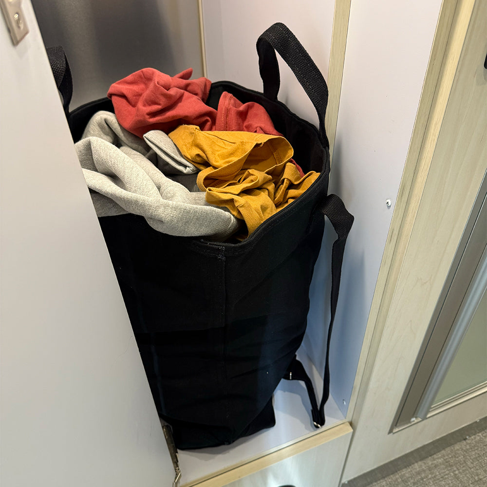 The Laundry Tote is a black, heavy-duty canvas bag filled with assorted red, gray, and yellow clothes, neatly placed inside a narrow closet or storage area. Designed for convenience, this bag features backpack-style straps for easy carrying.