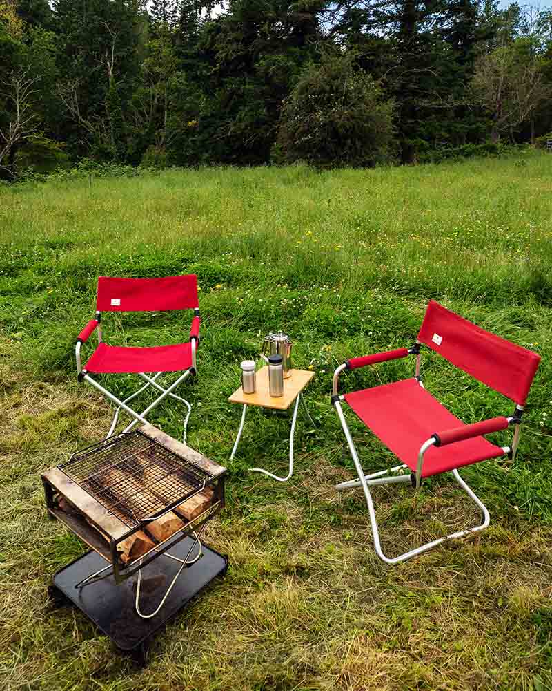 Two red folding chairs from the Snow Peak Red Lounge Set sit on grass beside a small table with mugs and a coffee pot. An empty, portable grill is set up nearby, surrounded by comfortable camping furniture. Trees and a meadow are in the background.