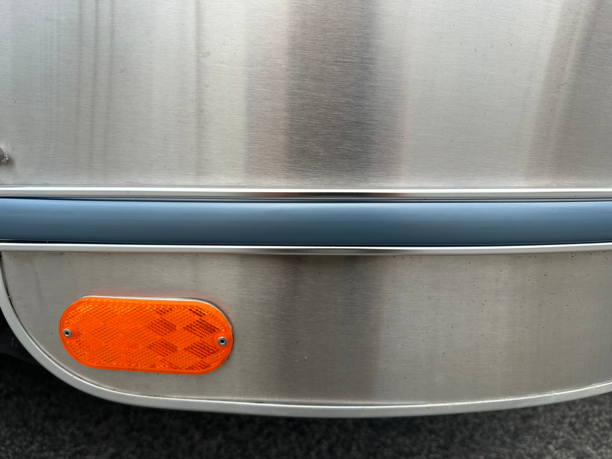 Close-up of the Airstream Premium Replacement Rub Rail for Travel Trailers, featuring a metallic surface with a blue horizontal stripe and an orange reflective marker near the bottom left corner. The smooth, slightly curved background highlights the distinctive riveted aluminum design characteristic of Airstream rub rails.