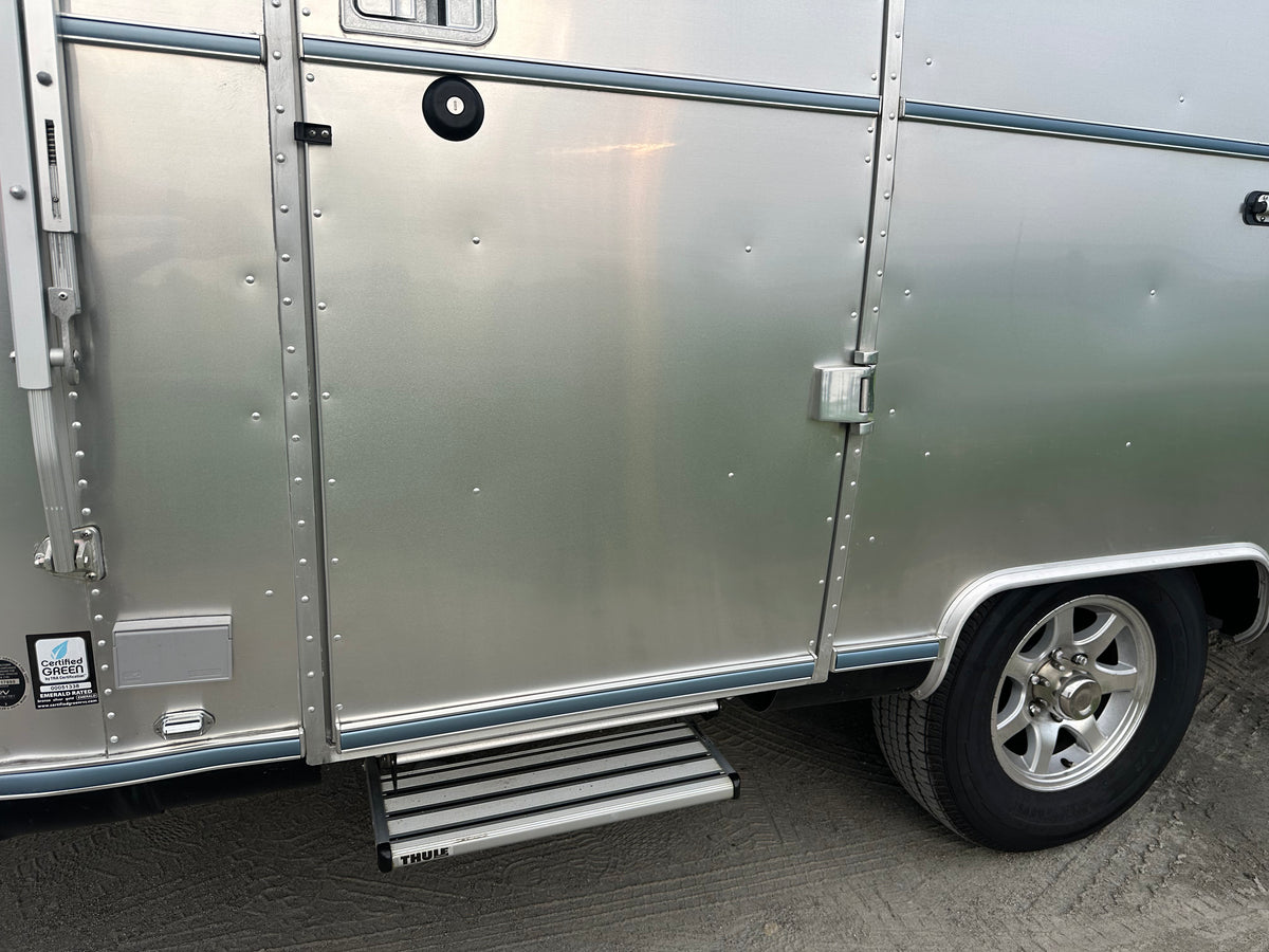 A close-up of an Airstream trailer door showcases the silver, riveted aluminum exterior, a retractable step, and a wheel on sandy ground, featuring the Airstream Premium Replacement Rub Rail for Travel Trailers.