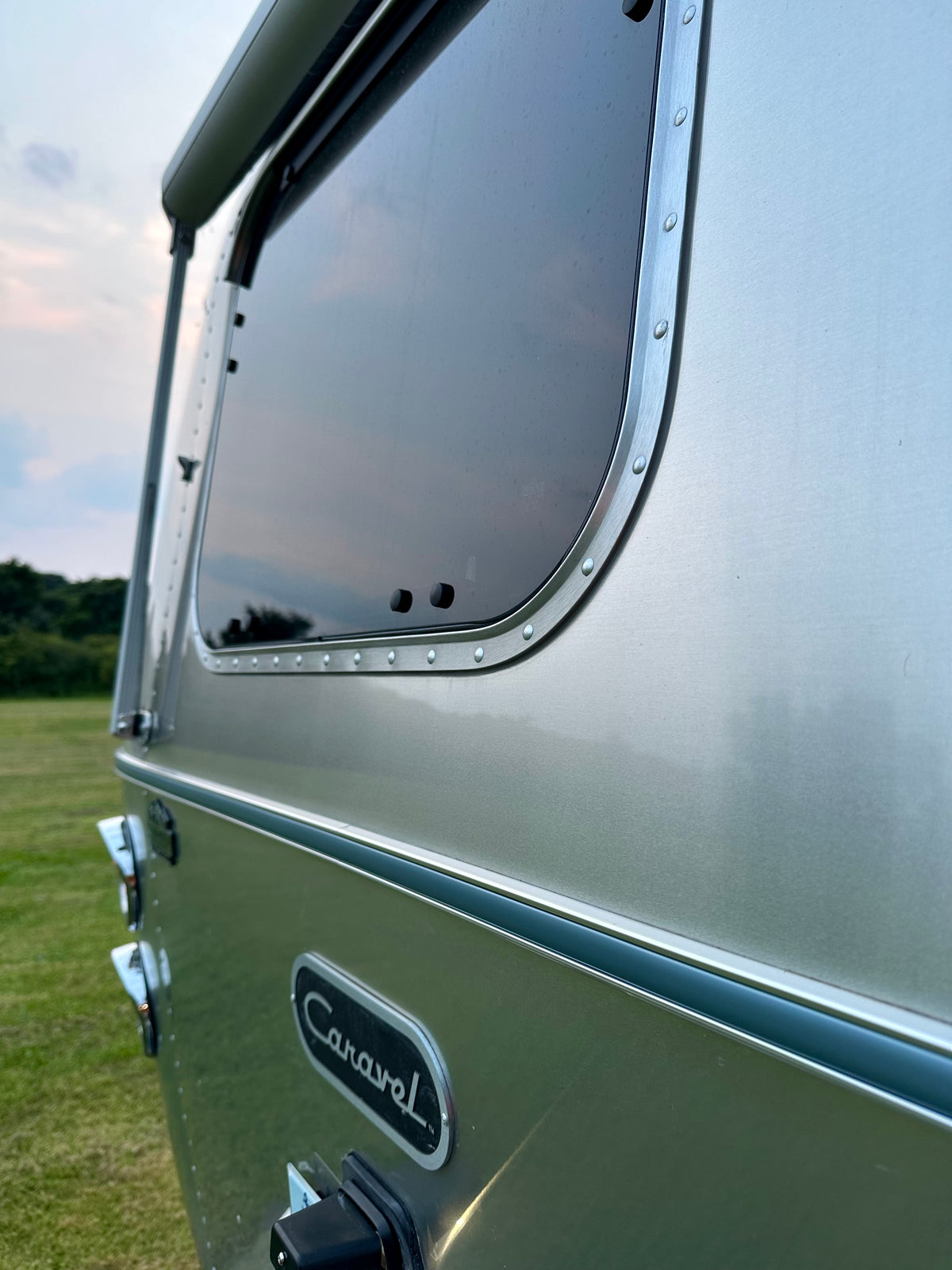 Close-up of a silver Airstream trailer featuring gleaming aluminum trim fitted with the Airstream Premium Replacement Belt Trim for Travel Trailers and a reflective window with the iconic Caravel badge, parked on grass with trees and a cloudy sky in the background.
