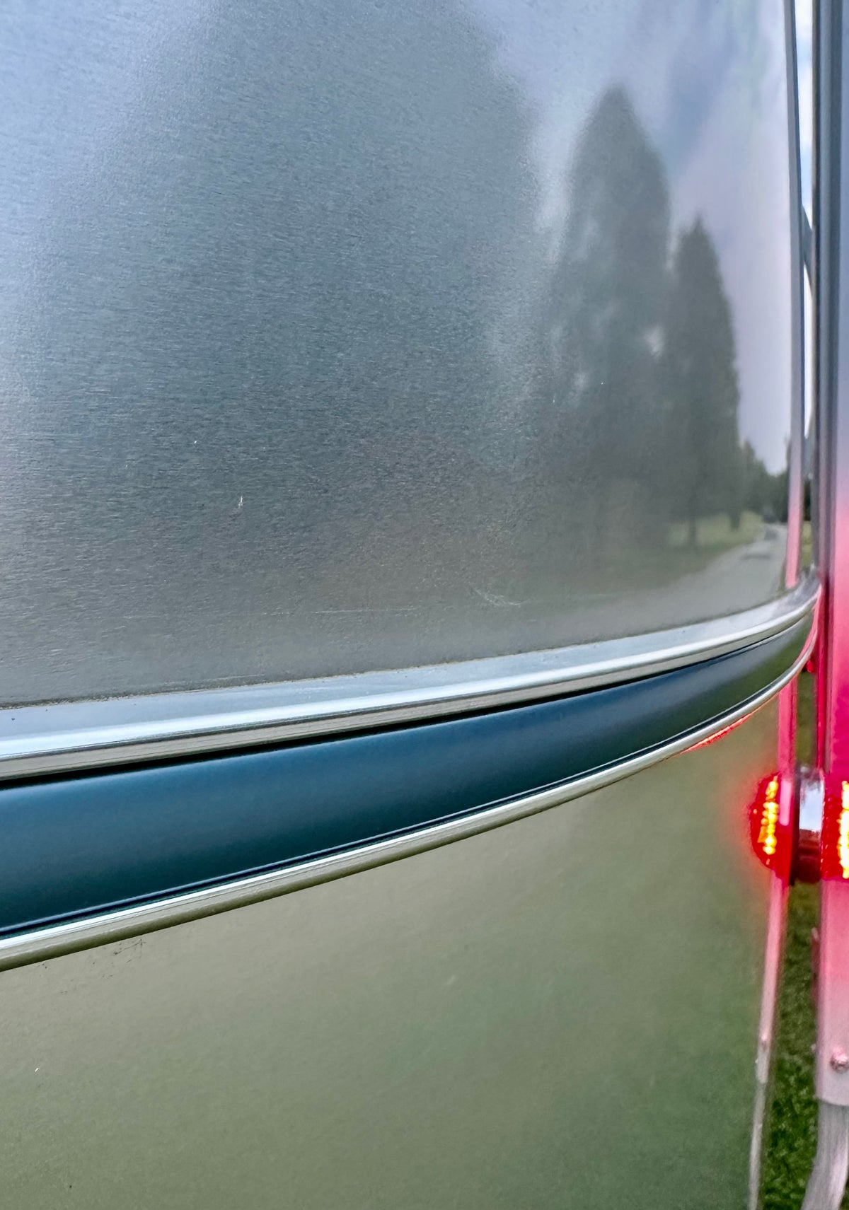 Close-up of a metallic trailer surface with a curved line, reflecting trees and sky. A red brake light is illuminated on the right, and grass is visible in the background. The polished aluminum Airstream Premium Replacement Belt Trim for Travel Trailers adds a sleek touch to the exterior accessory, enhancing both style and functionality.