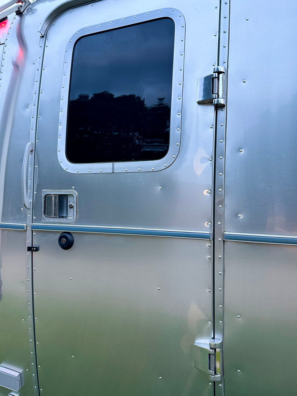 Close-up of a shiny silver trailer door showcasing the Airstream Premium Replacement Belt Trim for Travel Trailers, with a rectangular window reflecting trees, a sturdy handle, secure lock, and visible rivets along the sleek metal surface—an impressive example of an Airstream exterior accessory.