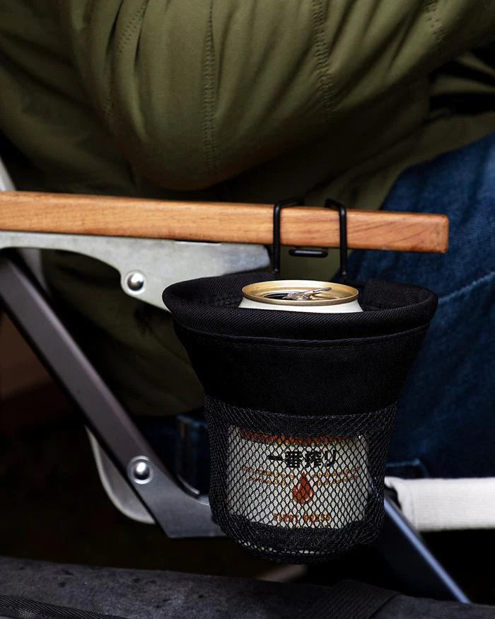 A person in a green jacket and blue jeans is sitting in a low beach chair equipped with the Snow Peak Low Chair Cup Holder for Low Beach Chair, which is attached to the wooden armrest and holds a can, offering a practical accessory for camping.