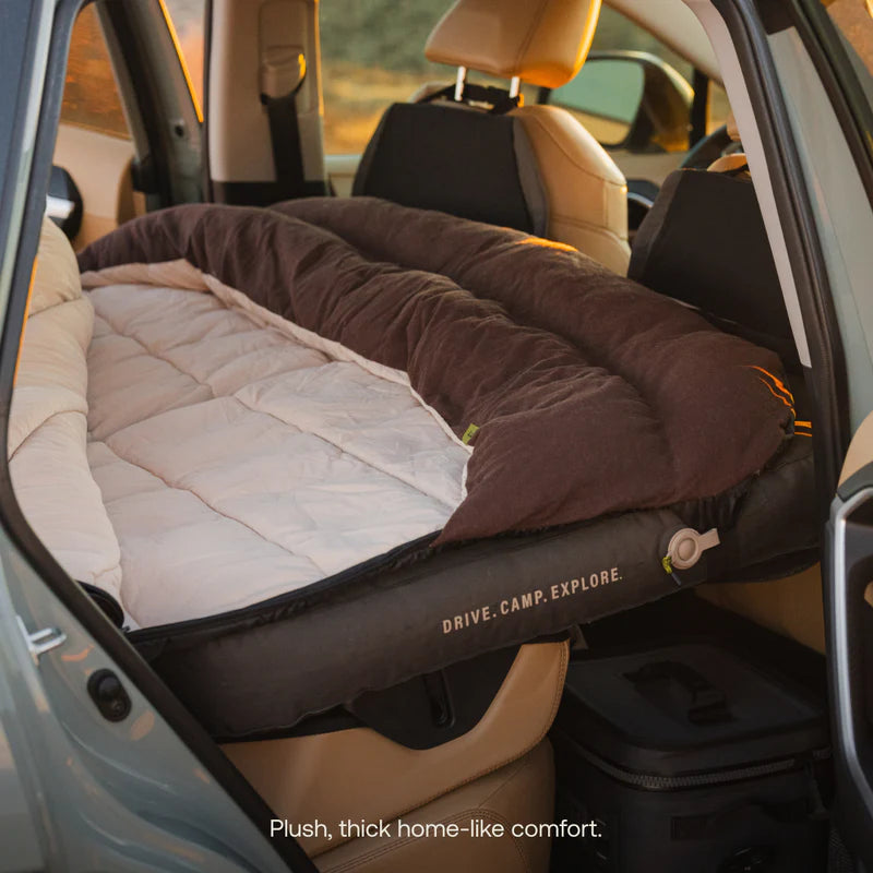 A thick, brown and beige Fitted Sleeping Bag Comforter by Luno is set up in the backseat of a car, creating a cozy bed reminiscent of an outdoor duvet. The words DRIVE. CAMP. EXPLORE. are printed on the side.