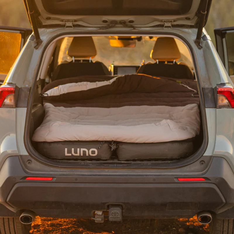 An SUV with its back hatch open displays a Luno air mattress in the cargo area, covered with bedding and topped with the Fitted Sleeping Bag Comforter by Luno, creating a cozy setup perfect for camping or sleeping outdoors.