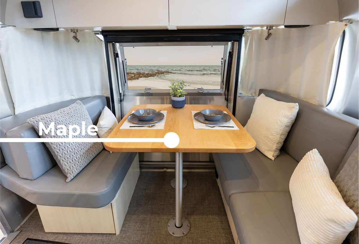 Inside an Airstream Flying Cloud Travel Trailer, a cozy dining nook showcases the Wood Dinette Table Upgrade, set for two with gray cushioned benches, decorative pillows, and a window framing a beach view. A label reads “Maple,” with a white line pointing to the table.