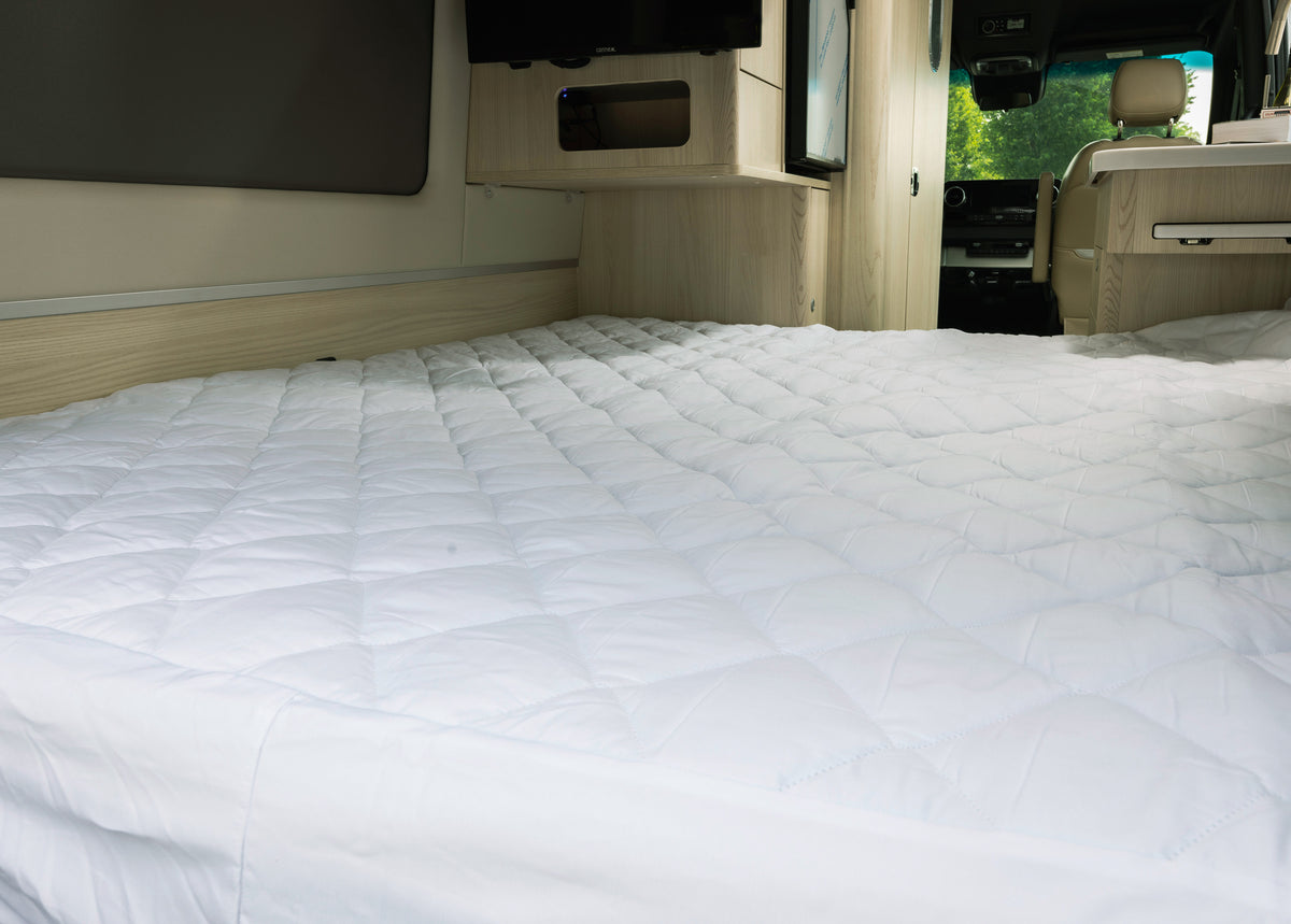 A neatly made bed featuring the Airstream Mattress Pad for Interstate 24 GL/GLX and GT/GTX—white, quilted, washable, and breathable—inside a modern camper van with light wood cabinetry and a window overlooking lush greenery.