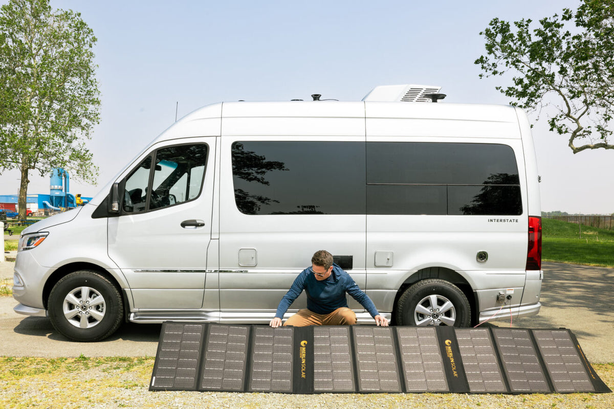 A man kneels in front of a silver Airstream Touring Coach, setting up the Merlin Solar Panel Bundle for Airstream Touring Coaches on the ground next to the vehicle. Trees and a clear sky are visible in the background.