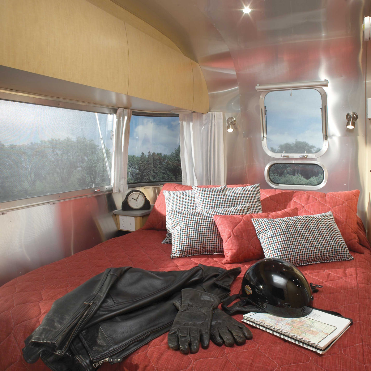 A cozy trailer bedroom features a red bedspread, patterned pillows, a leather jacket, gloves, helmet, and notebook on the bed. Airstream Solid Custom Curtains for Pan America Travel Trailers frame the window, where a clock sits nearby and trees are visible outside.