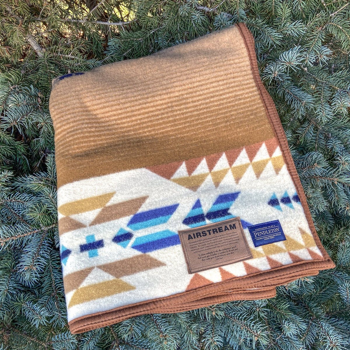 A folded Airstream x Pendleton Woolen Mills Jacquard Blanket with a geometric pattern in tan, white, blue, and brown rests on green pine needles. The blanket features labels reading AIRSTREAM and PENDLETON attached to one corner.