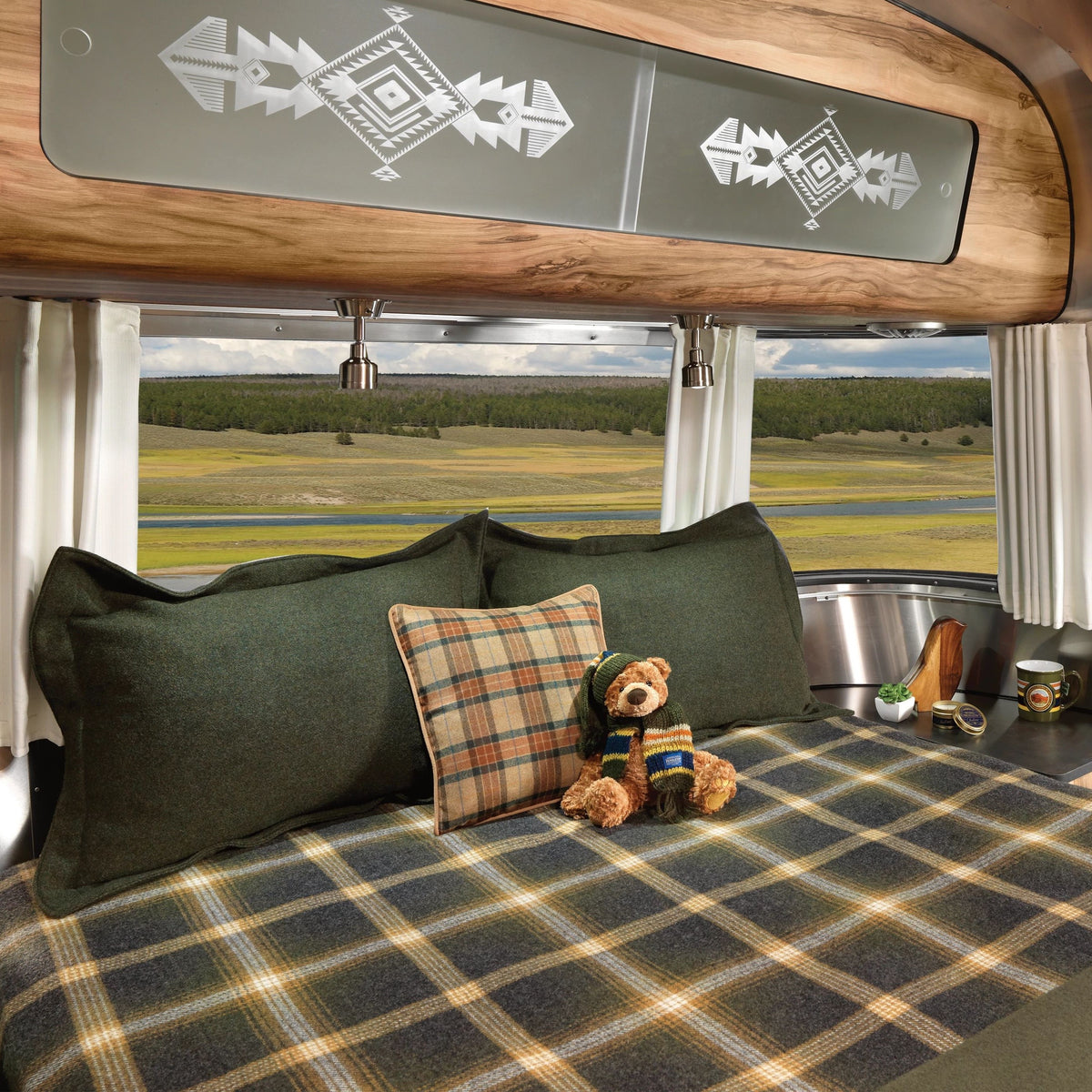 A cozy bed with green plaid bedding, a matching plaid pillow, and a stuffed bear sits inside a trailer featuring Airstream Solid Custom Curtains for Pendleton Travel Trailers framing the window, showcasing fields and trees outside.