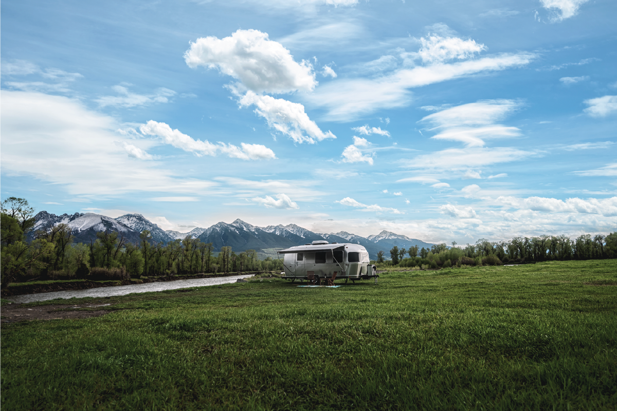 An Airstream Travel Trailer from the “Picturesque Postcards” collection is parked on a grassy field by a river, set against a backdrop of mountains and trees beneath a blue, cloud-filled sky—capturing the timeless charm of scenic postcards.
