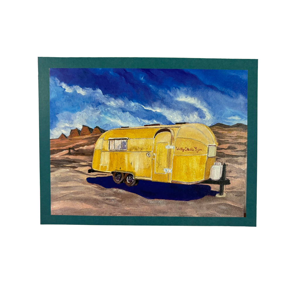 A painting of a yellow vintage trailer on a desert landscape beneath a dramatic blue sky with clouds and distant rock formations, bordered by a dark green mat—an ideal addition to your travel greeting cards or the Airstream Premium Card Gift Set.