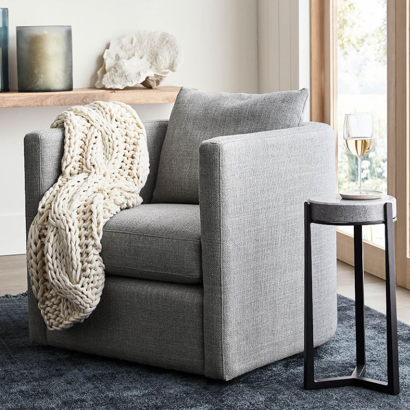 A modern gray armchair draped with the Airstream x Pottery Barn Colossal Handknit Throw and a matching cushion sits on a dark rug. Beside it, a small round side table holds a glass of white wine, with sunlight streaming in through large windows.