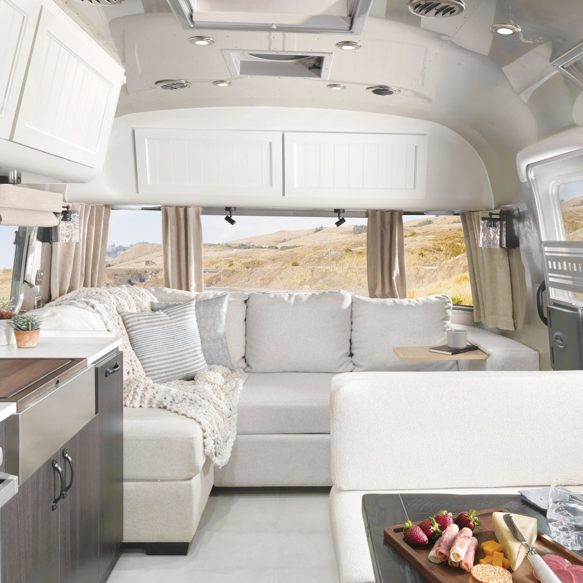 Modern RV interior with a light gray sectional sofa, cozy blanket, small table, and large windows revealing a scenic landscape. Freshly installed Airstream Solid Custom Curtains for Pottery Barn Travel Trailers frame the windows, complementing the soft décor. A countertop and charcuterie board are visible in the foreground.