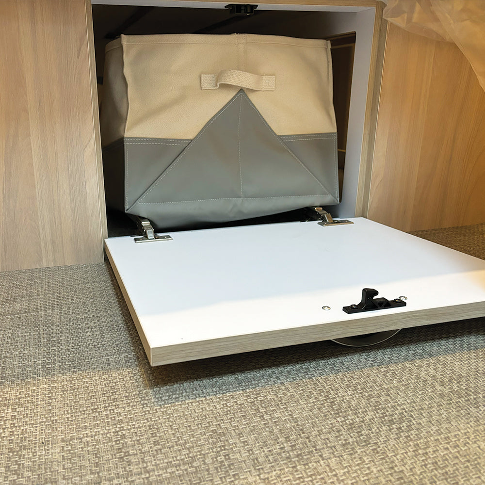 An Airstream x Steele Canvas Queen Bed Storage Bin, featuring a gray and beige heavy-duty canvas design, is placed inside a wooden cubby with an open white pull-out door, resting on a beige textured carpet.