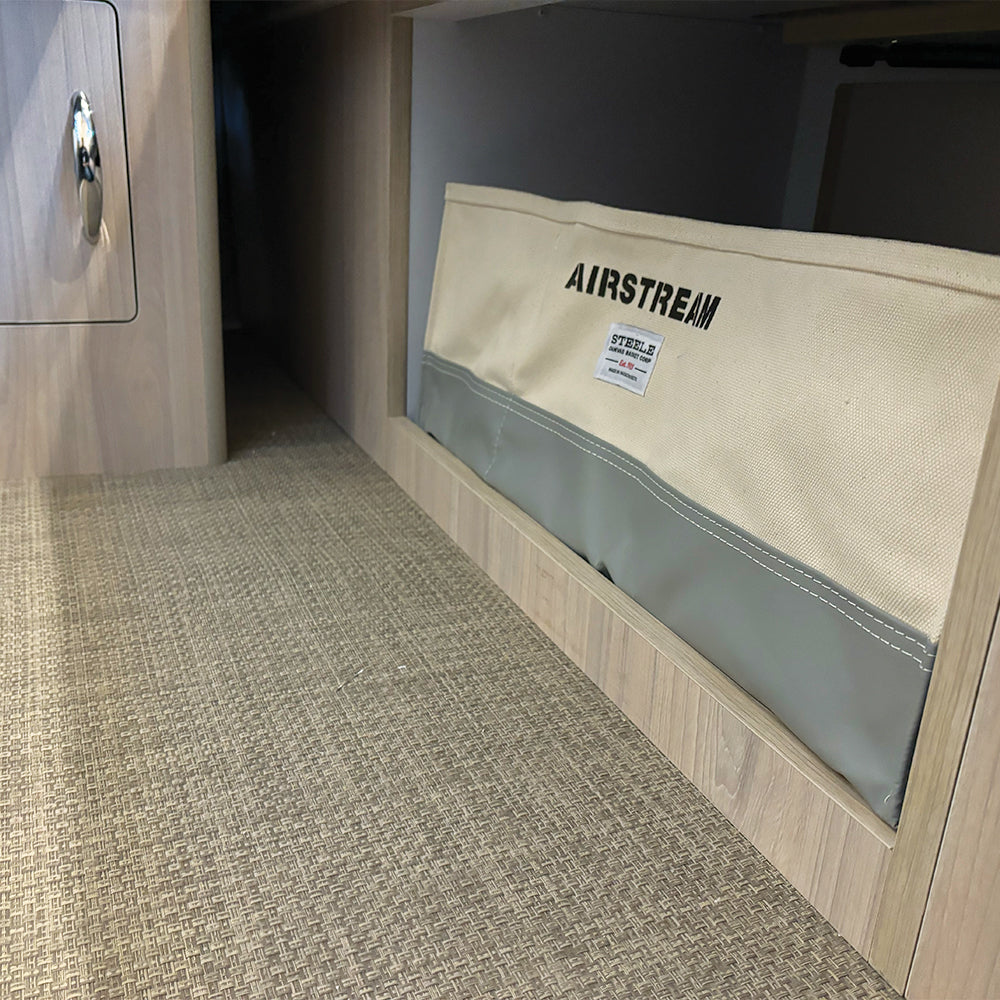 An Airstream x Steele Canvas Queen Bed Storage Bin in beige and gray is built into a wood-paneled wall, accompanied by textured brown flooring and a nearby queen bed floor cabinet.