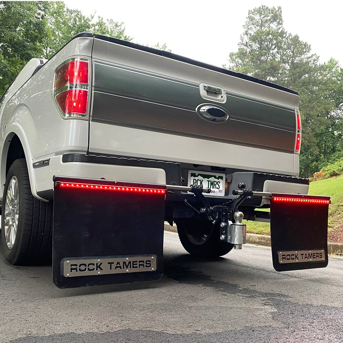 Rear view of a silver pickup truck equipped with large black mud flaps labeled Rock Tamers and featuring Rock Tamers Tail Light Bars with red LED trailer lights, parked on a paved surface with trees and grass in the background. A 4-wire flat trailer connector is visible beneath the bumper, enhancing its towing capabilities.
