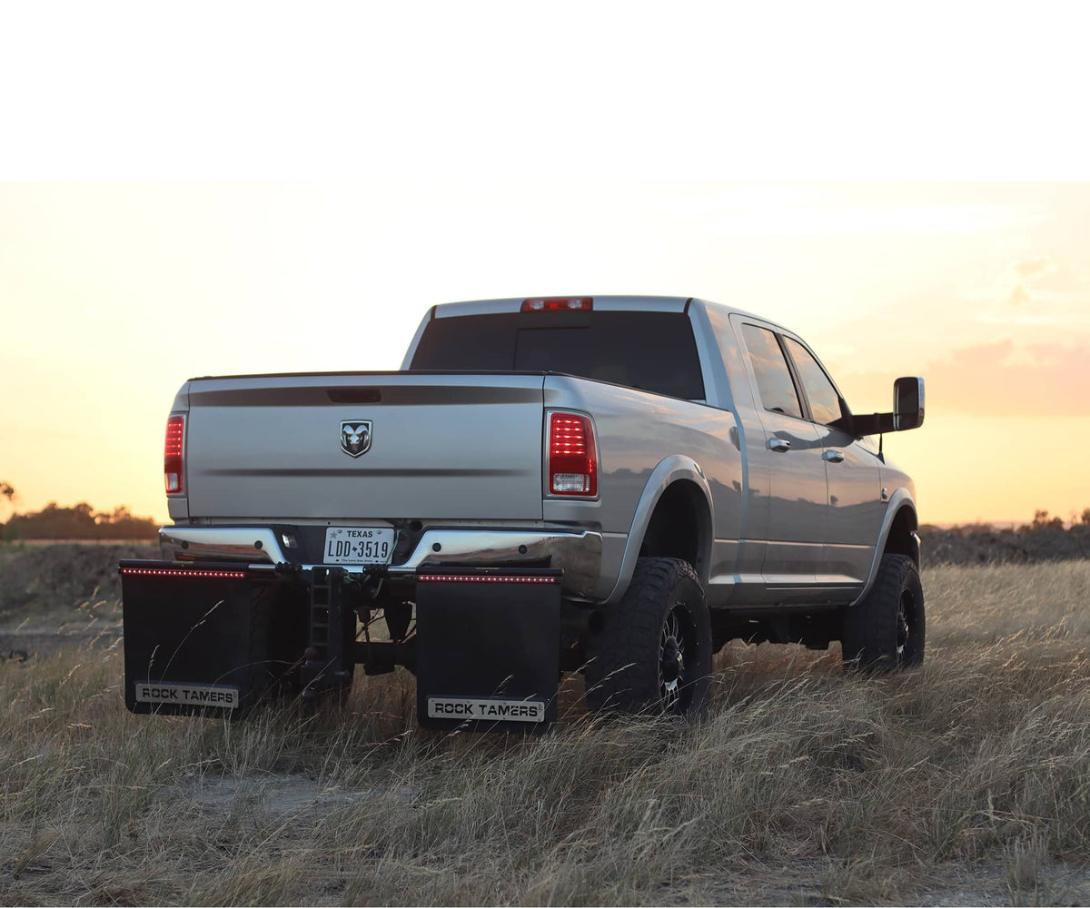 A silver Ram pickup truck with large tires, Rock Tamers Tail Light Bars, and mud flaps is parked in a grassy field at sunset.