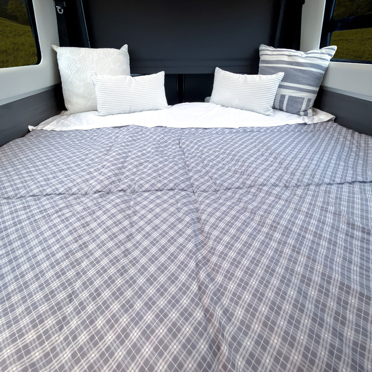 Inside a vehicle with windows, a neatly made bed features the Airstream x RVSuperbag All-In-One Bedding System—a gray plaid comforter paired with four decorative pillows—creating a cozy and inviting sleeping space perfect for your Airstream adventures.