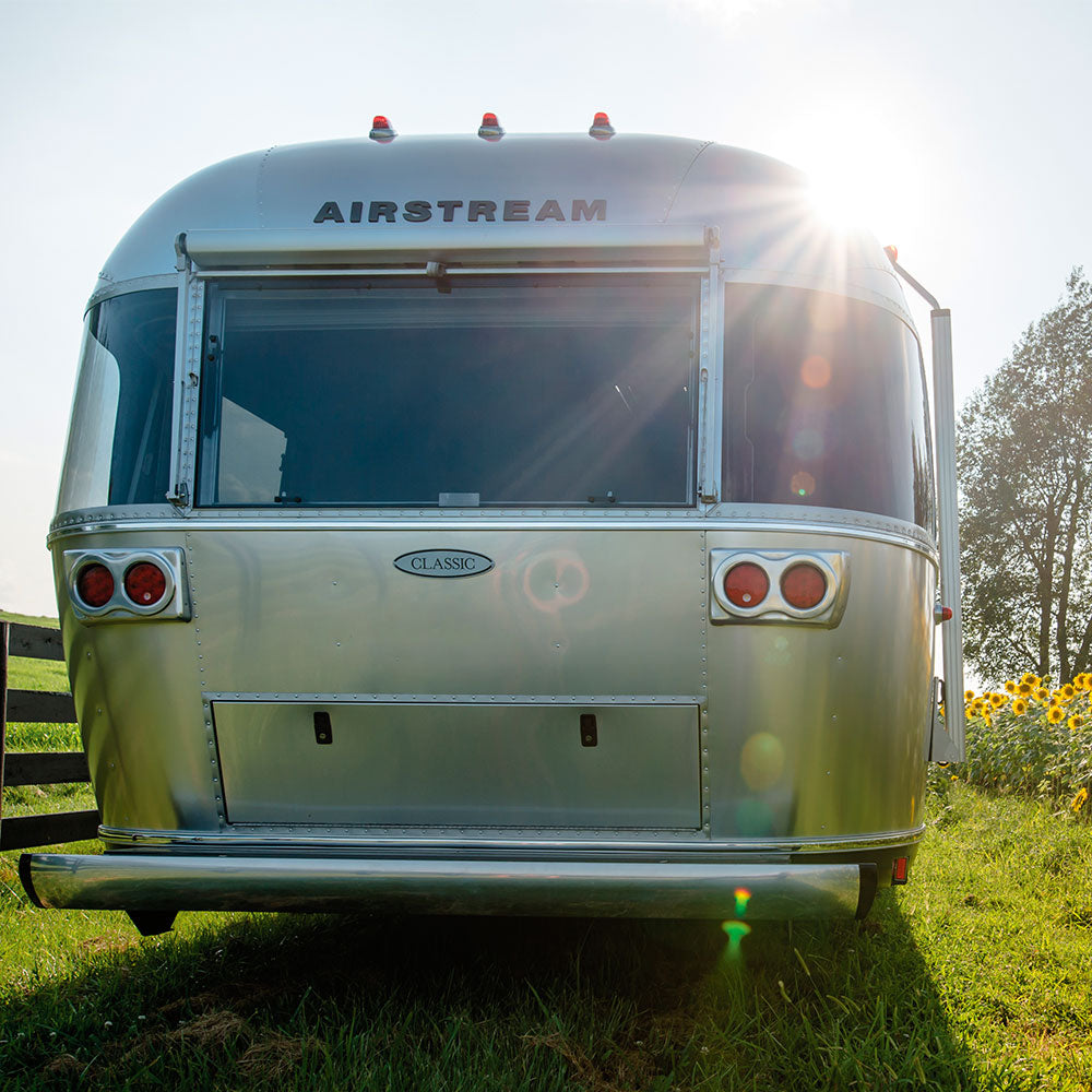 A riveted aluminum Airstream trailer is parked on the grass, sunlight illuminating its iconic curves and shimmering exterior. To the right, sunflowers and trees highlight the distinctive Airstream Premium Replacement Rub Rail for Travel Trailers installed along its side.