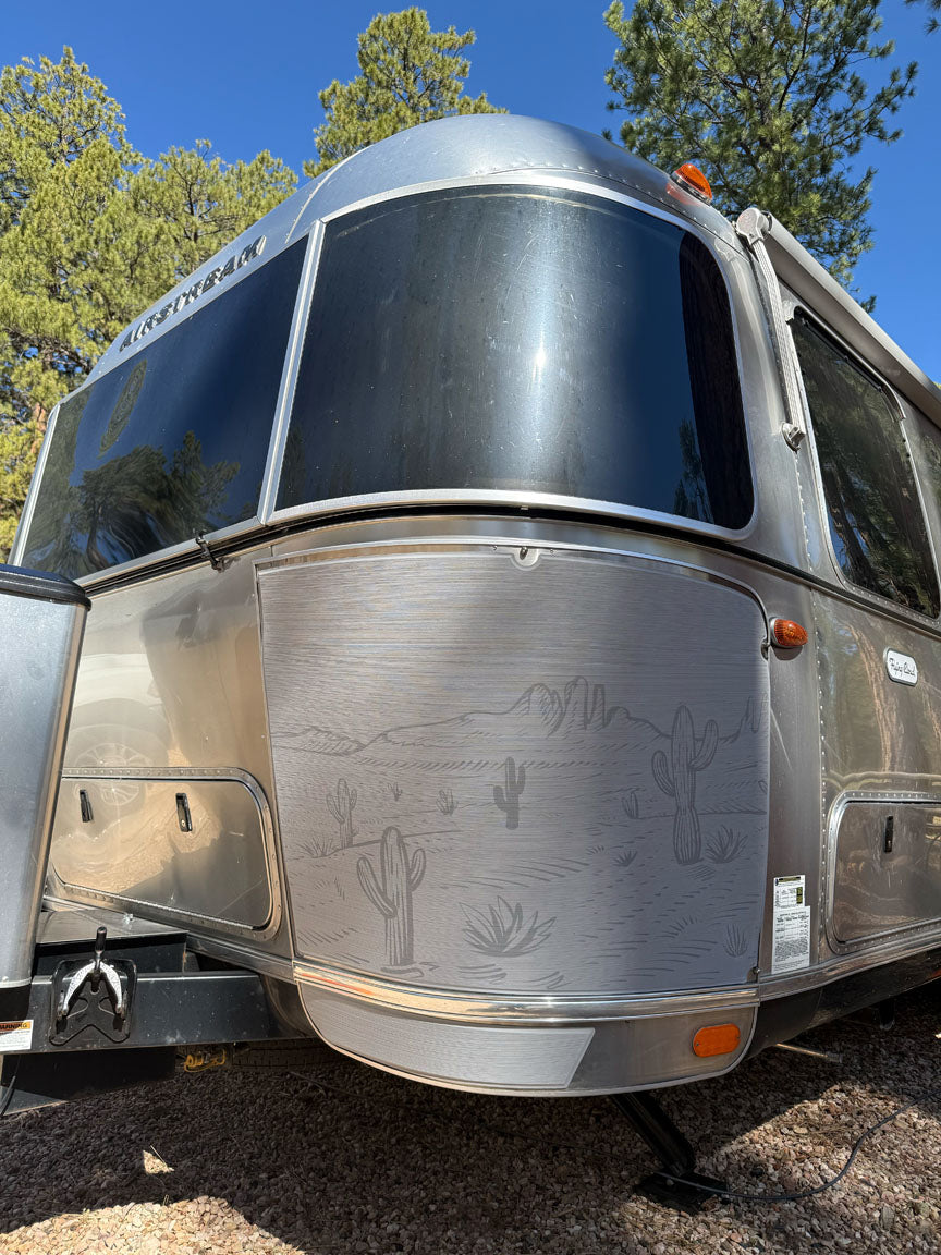 A close-up of a silver Airstream trailer parked outdoors among tall pine trees under a clear blue sky, featuring desert cactus designs etched on the front and equipped with Airstream Rock Guard Rock Wraps Nature Collection for added travel trailer protection.
