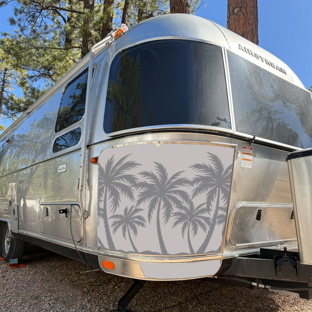 A silver Airstream trailer is parked outdoors among tall pine trees. The trailer features a gray decorative cover from the Airstream Rock Guard Rock Wraps Nature Collection, showcasing a palm tree silhouette design on its front for added protection and style while traveling.