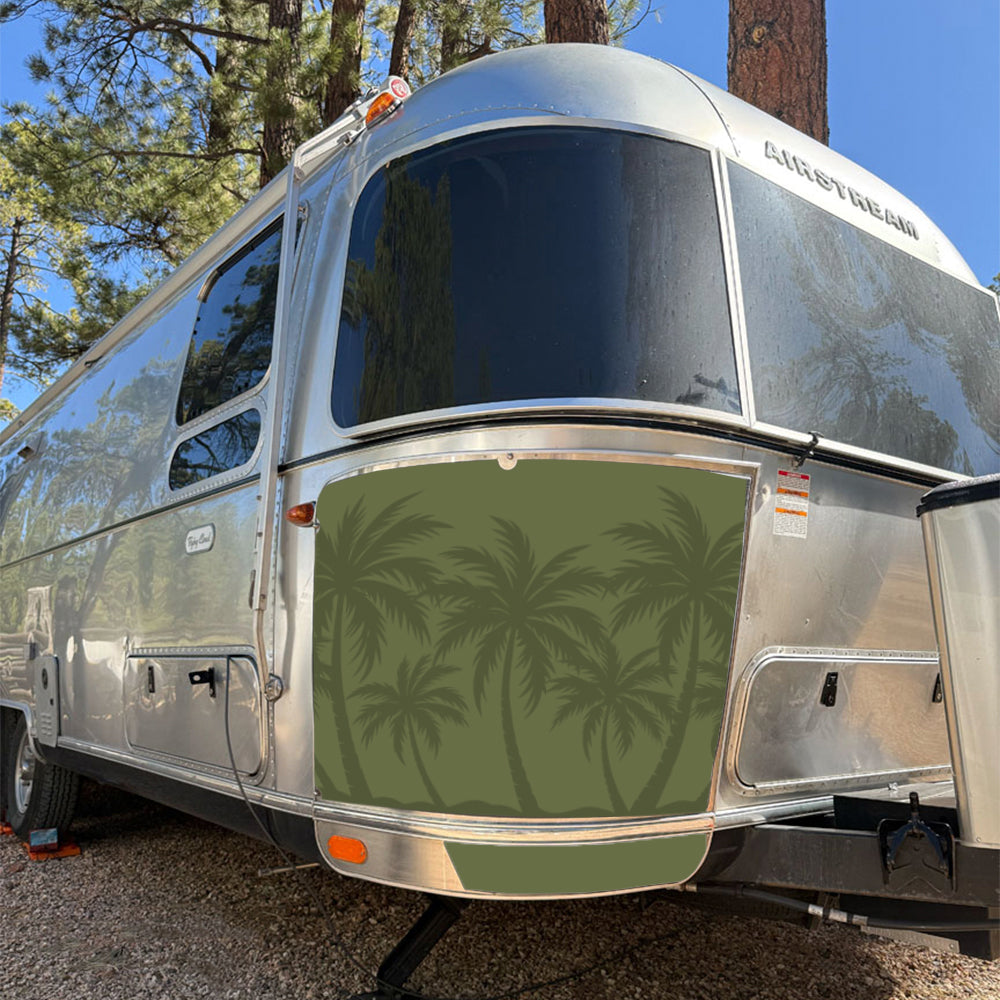 A silver Airstream trailer is parked outdoors among tall trees. The front lower panel displays a green cover with palm tree silhouettes, featuring the Airstream Rock Guard Rock Wraps Nature Collection for added travel trailer protection.