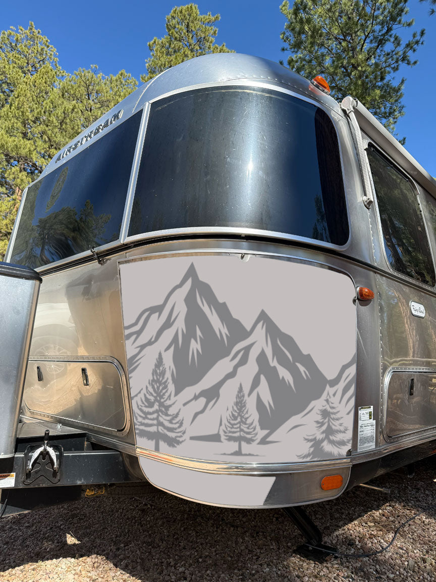 An Airstream trailer featuring a gray mountain and tree design on its front is parked outdoors among pine trees under a sunny sky, with its exterior protected by durable Airstream Rock Guard Rock Wraps from the Nature Collection.
