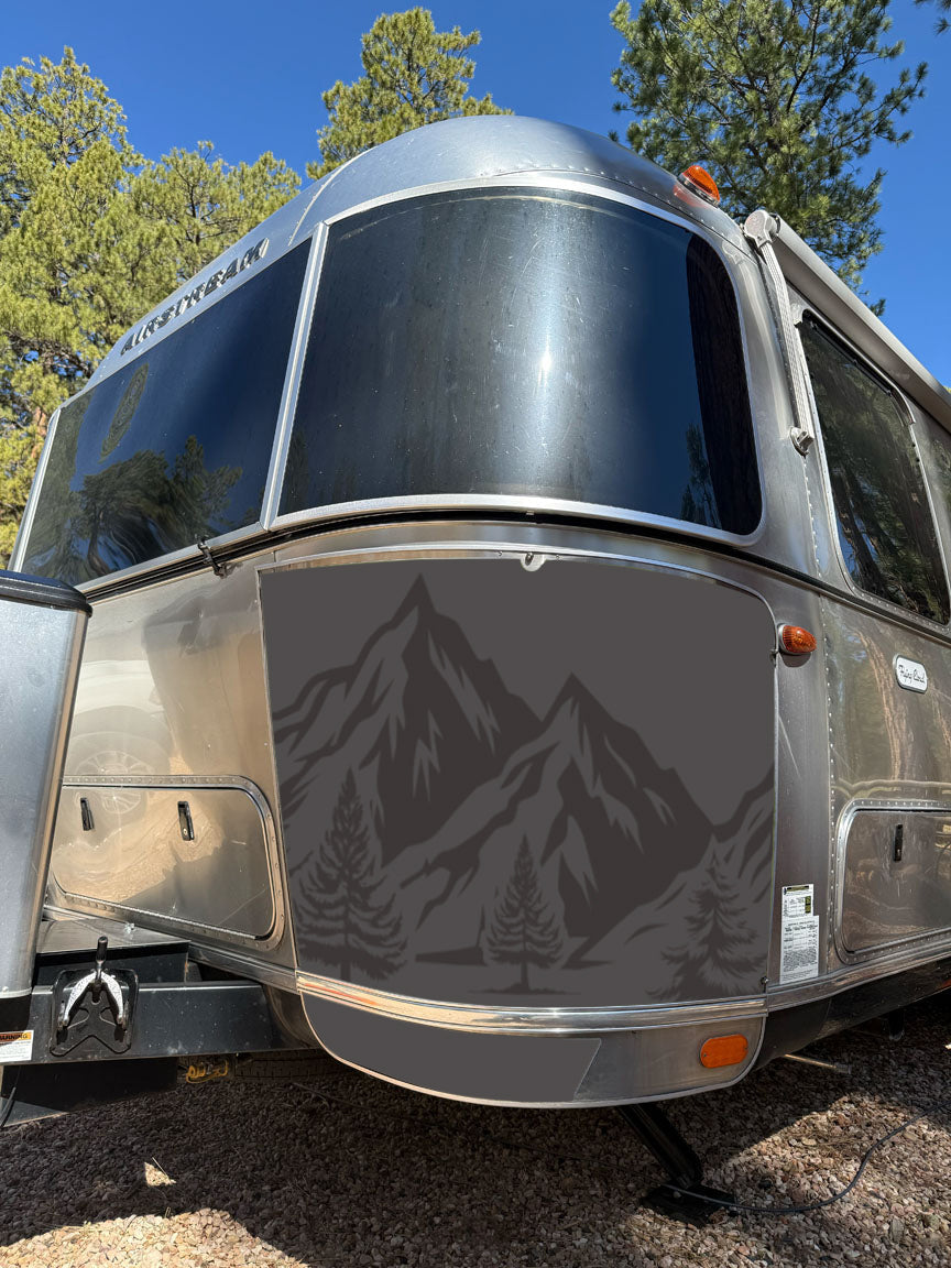 A silver Airstream trailer is parked outside, adorned with a dark mountain and tree decal on the front panel, framed by tall pine trees and a blue sky in the background. The front features the durable Airstream Rock Guard Rock Wraps Nature Collection, providing enhanced protection for your travel adventures.