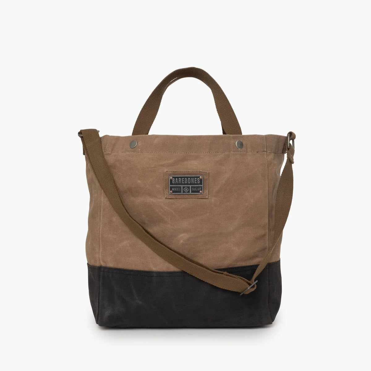 The Neelum Tote Bag by Barebones is a durable and stylish two-tone waxed canvas tote featuring a brown upper, black bottom section, top handle, adjustable shoulder strap, and front patch label—making it an ideal reusable bag for everyday use.